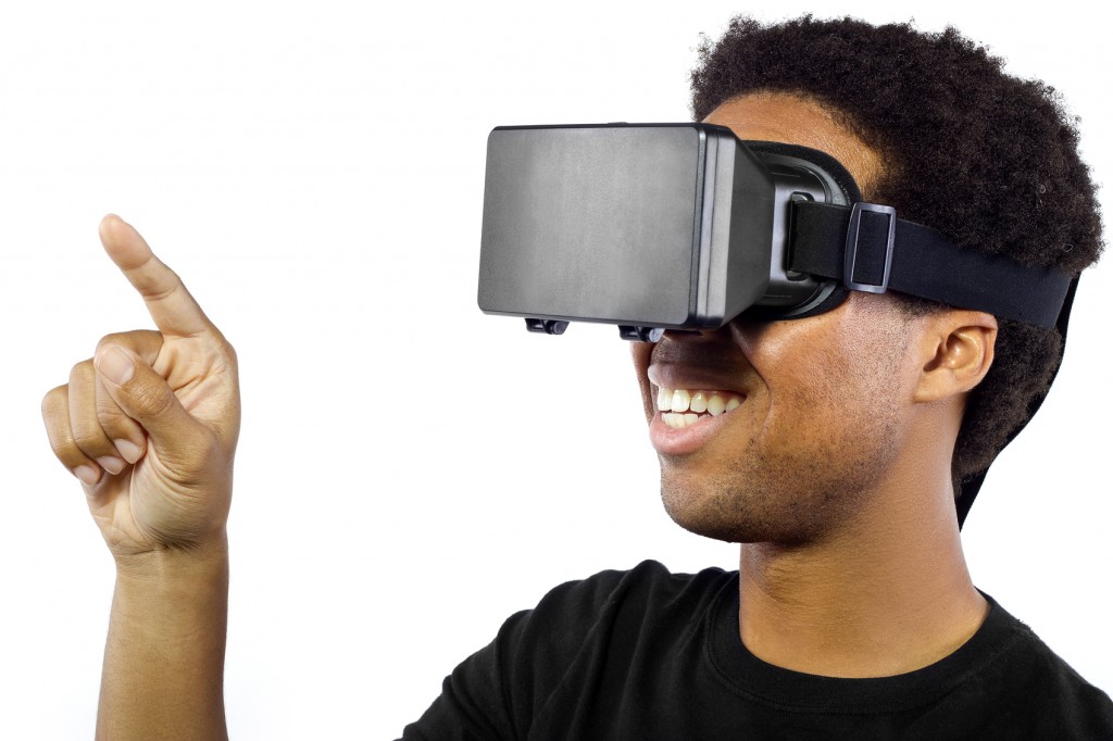 Photo of a black male wearing a virtual reality headset on a white background. The headset is worn over the eyes as goggles and has a digital screen. The device is for immersive VR experience. The image depicts innovation is video game VR technology. The man is pointing with his finger like he is interacting with something.