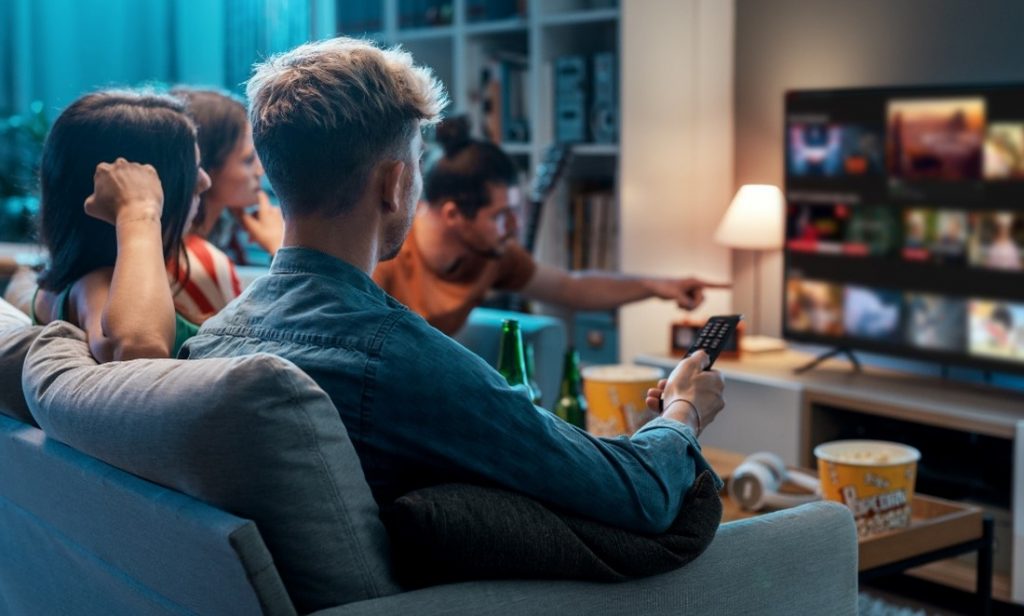 A group of people, 2 women and 2 men, are set on a sofa and watching tv