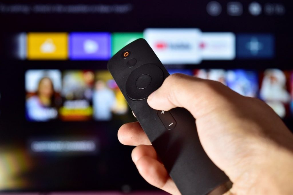 A simple black remote is pointed towards a large television screen. The screen has blurred images, the selection screen has various streaming services listed. 